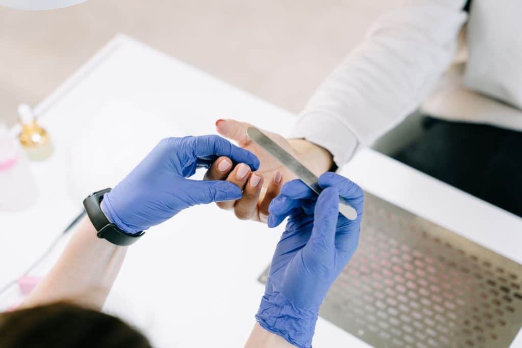 Nail technician in blue gloves filing a client's nail to prep a lifted gel patch